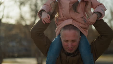 a man wearing a brown jacket carries a little girl in a pink cap and jacket on his neck, creating a joyful and playful moment outdoors in a park setting with trees and sunlight in the background