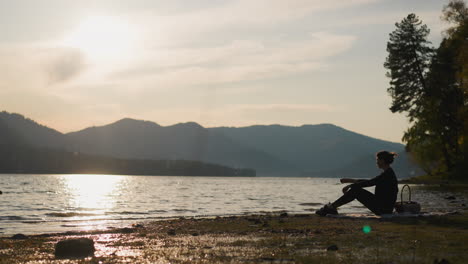 dama arrojando piedras de guijarros al lago. mujer con canasta de comida pasa el tiempo disfrutando de un momento de tranquilidad en la orilla del río al atardecer. fin de semana sola