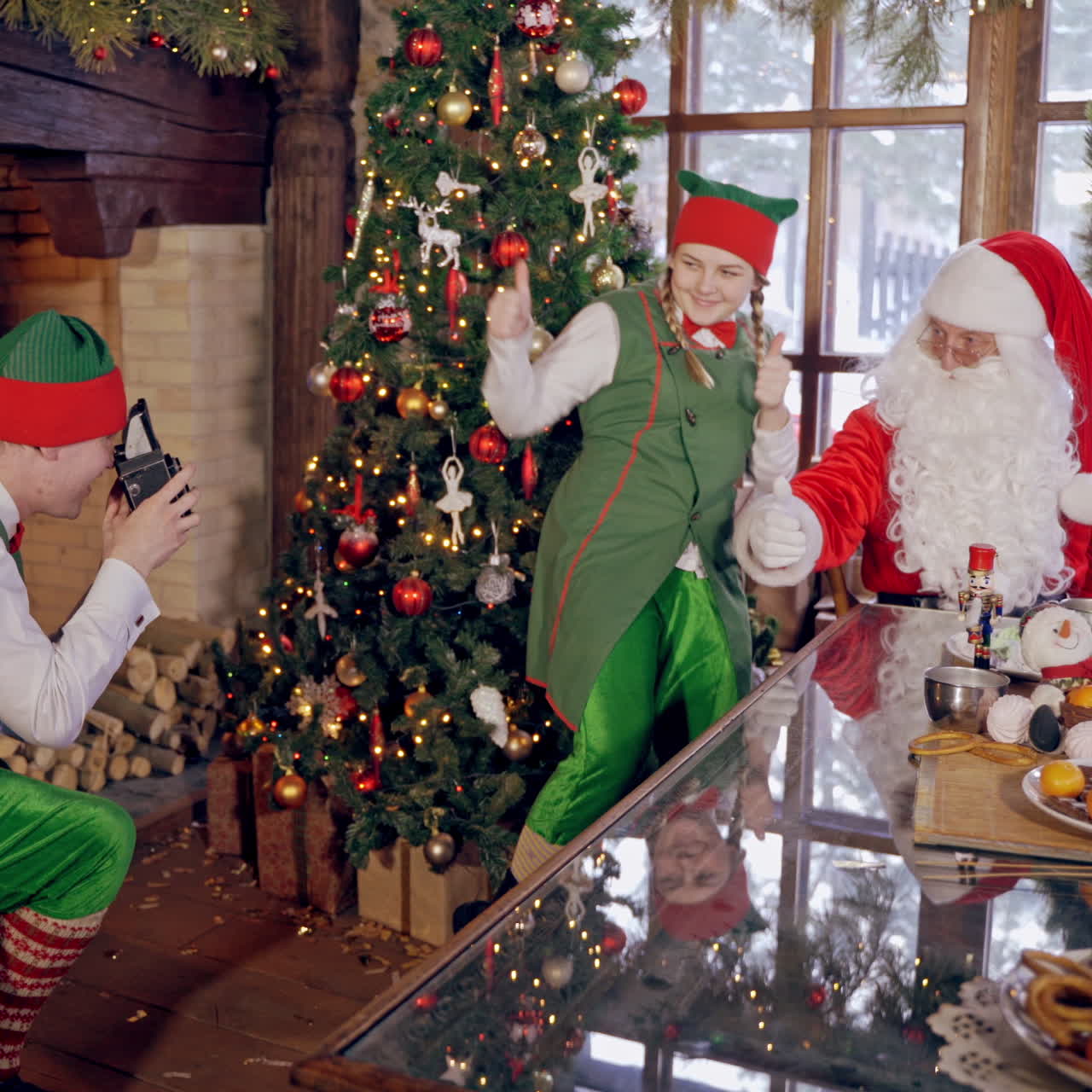 Santa together with elf near the Christmas tree. Funny elf is taking photo of fairy characters in Christmas decorated room.