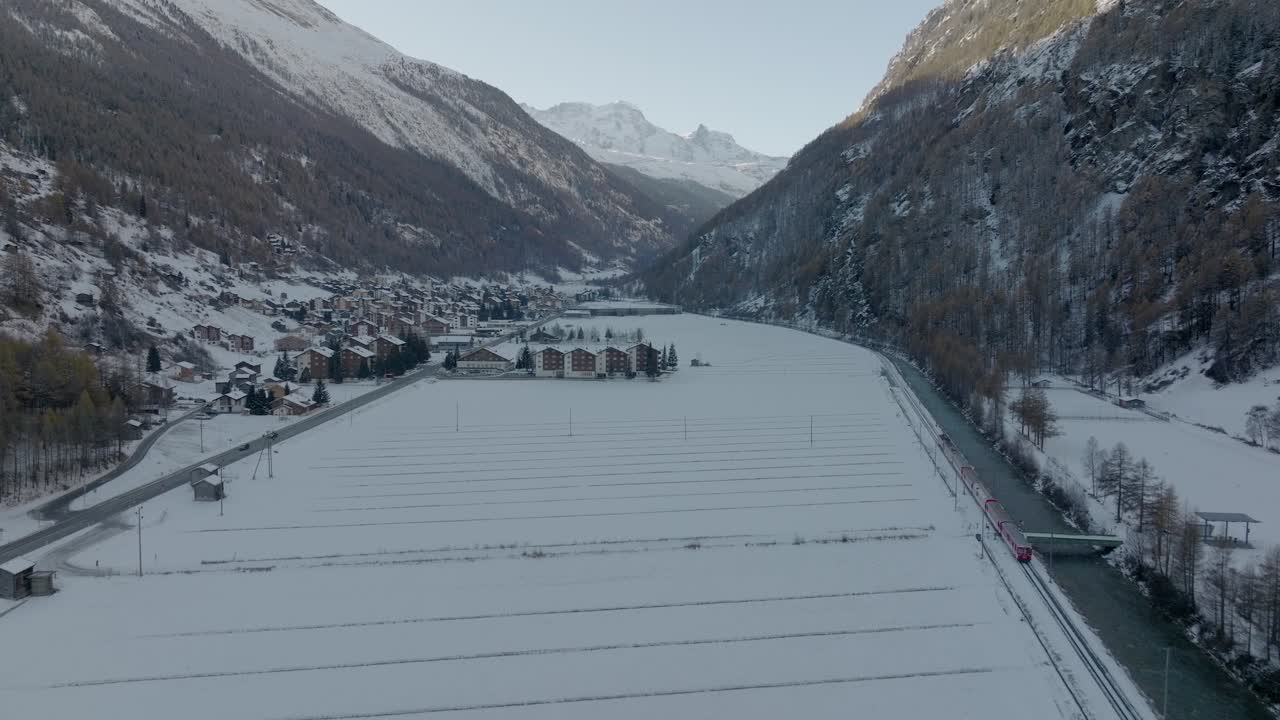 toma aérea de un tren rojo en suiza recorriendo un impresionante paisaje nevado de invierno junto al río que fluye con panorama de montañas alpinas y un pequeño y lindo pueblo de montaña suizo