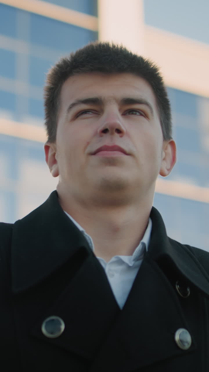 검은색 슈트를 입은 자신감 넘치는 남자가 자신감 넘친 표정으로 목걸이를 조정하며 우아함과 스타일을 보여줍니다. 배경에는 태양빛을 반사하는 현대적인 유리 건물이 있습니다.