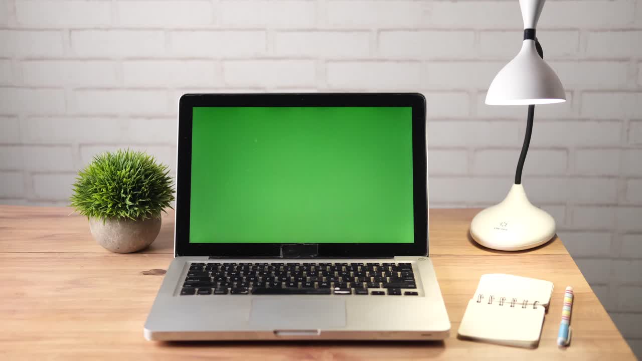 Laptop on a desk with green screen, lamp, plant, and notebook