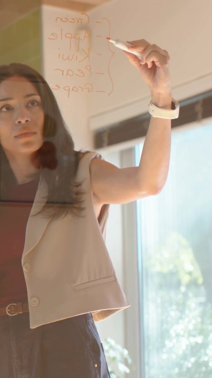 Video verticale: Alzando un pennarello, donna in gilet che elenca su vetro in ufficio vicino a una pianificazione con sbavature.