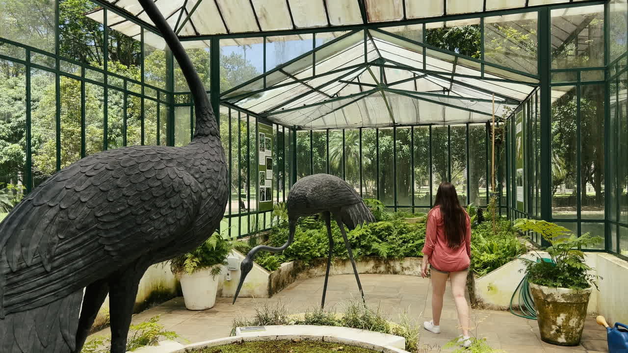 A woman walks among giant black crane sculptures and lush tropical plants inside the historic iron and glass Fern Greenhouse (Estufa de Samambaias) at the Rio de Janeiro Botanical Garden, Brazil