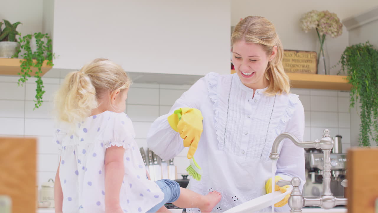 Mother wearing rubber gloves at home in kitchen with young daughter having fun and washing girl's feet as they do washing up at sink- shot in slow motion