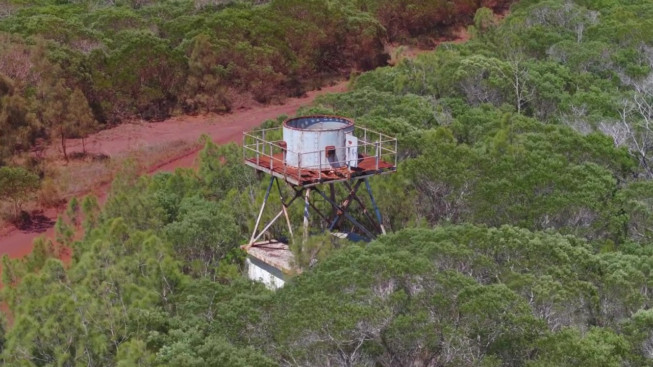 Drone approach over New Caledonia bushland toward the abandoned Tiébaghi precipitation radar, a damaged hilltop structure near a red dirt road in a remote landscape.