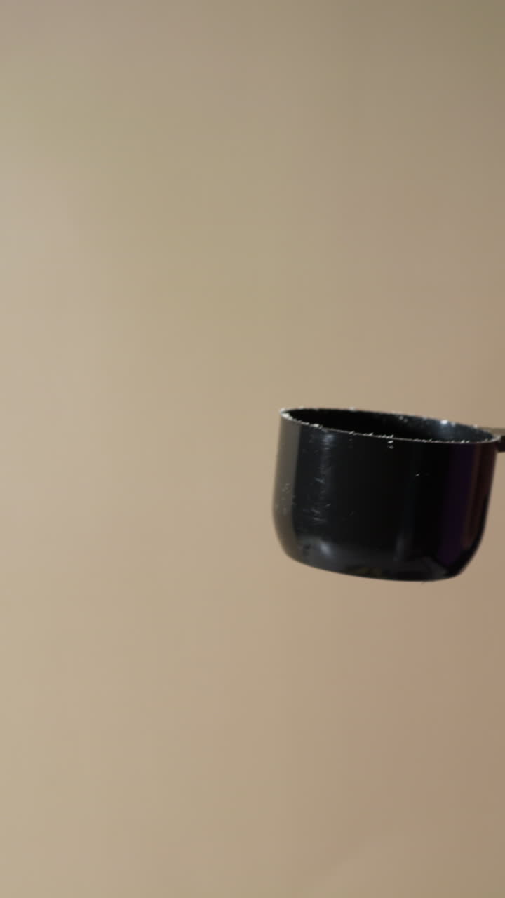 A close-up shot of protein powder gently tipping out of a black scoop held by hand, as fine particles fall in slow motion against a neutral beige background, suggesting vanilla or banana flavor.
