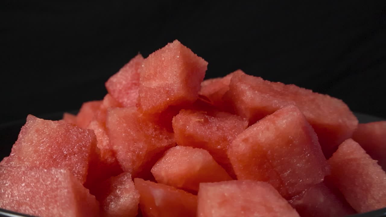 Close up video of fresh and tasty red colored watermelon sliced cubes in a bowl while placed in a pile in front of a black and dark blurry bokeh background. Juicy fruit ready to be eaten and snacked