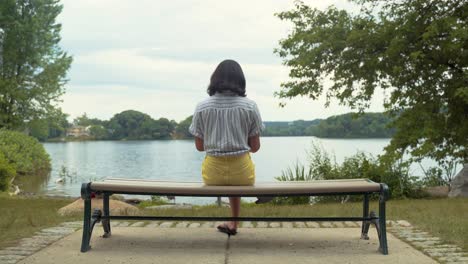 female student reading book on park bench by calm, peaceful lake, wide shot, filmed from behind