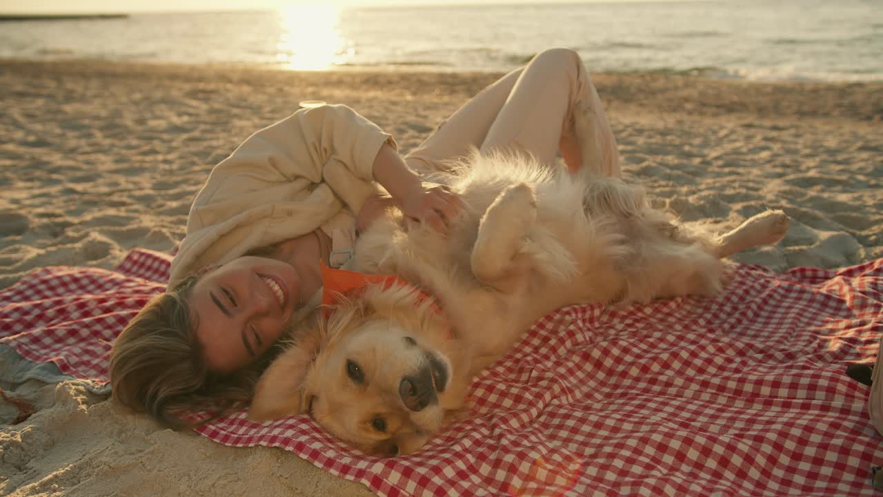 Close-up shot: a blonde girl with her light-colored dog lie on a mat on a sunny beach in the morning. The girl strokes the dog and she is happy