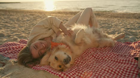 close-up shot: a blonde girl with her light-colored dog lie on a mat on a sunny beach in the morning. the girl strokes the dog and she is happy