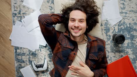 Portrait-of-a-happy-brunette-guy-in-a-plaid-shirt-who-lies-on-a-pillow-on-the-floor-among-sheets-of-papers-coffee-laptop-and-joystick-at-home