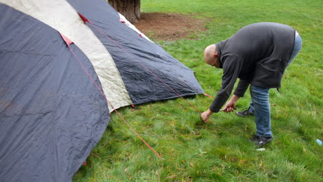 a mature man hammering a tent peg into the grass in a field at a campsite