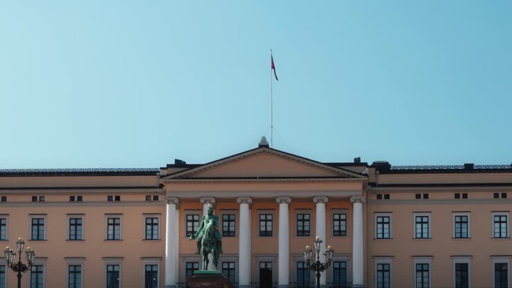 Palazzo Reale di Oslo, Norvegia, maestosa facciata gialla sotto un cielo azzurro limpido, con la statua equestre in bronzo del Re Karl Johan III e la bandiera norvegese in cima all'asta.