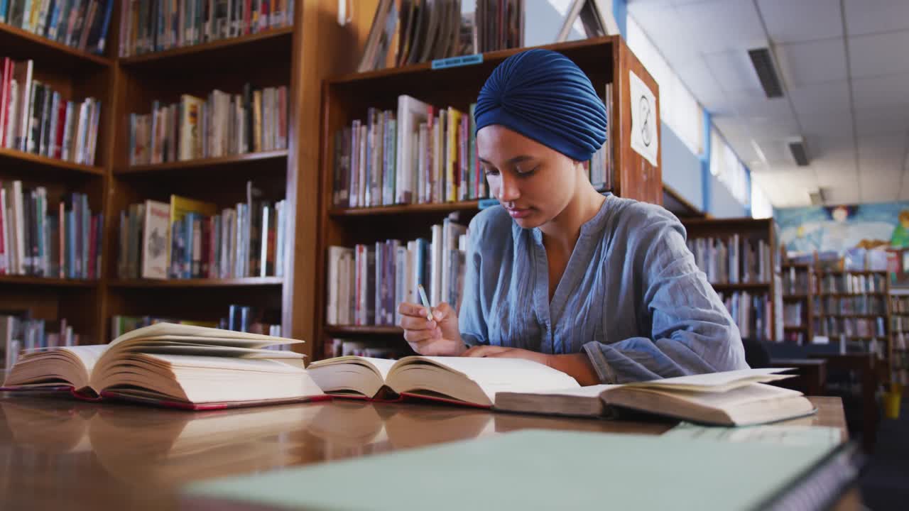 Asiatisk kvinnelig student med blå hijab sitter ved et skrivebord med åpne bøker og tar notater.