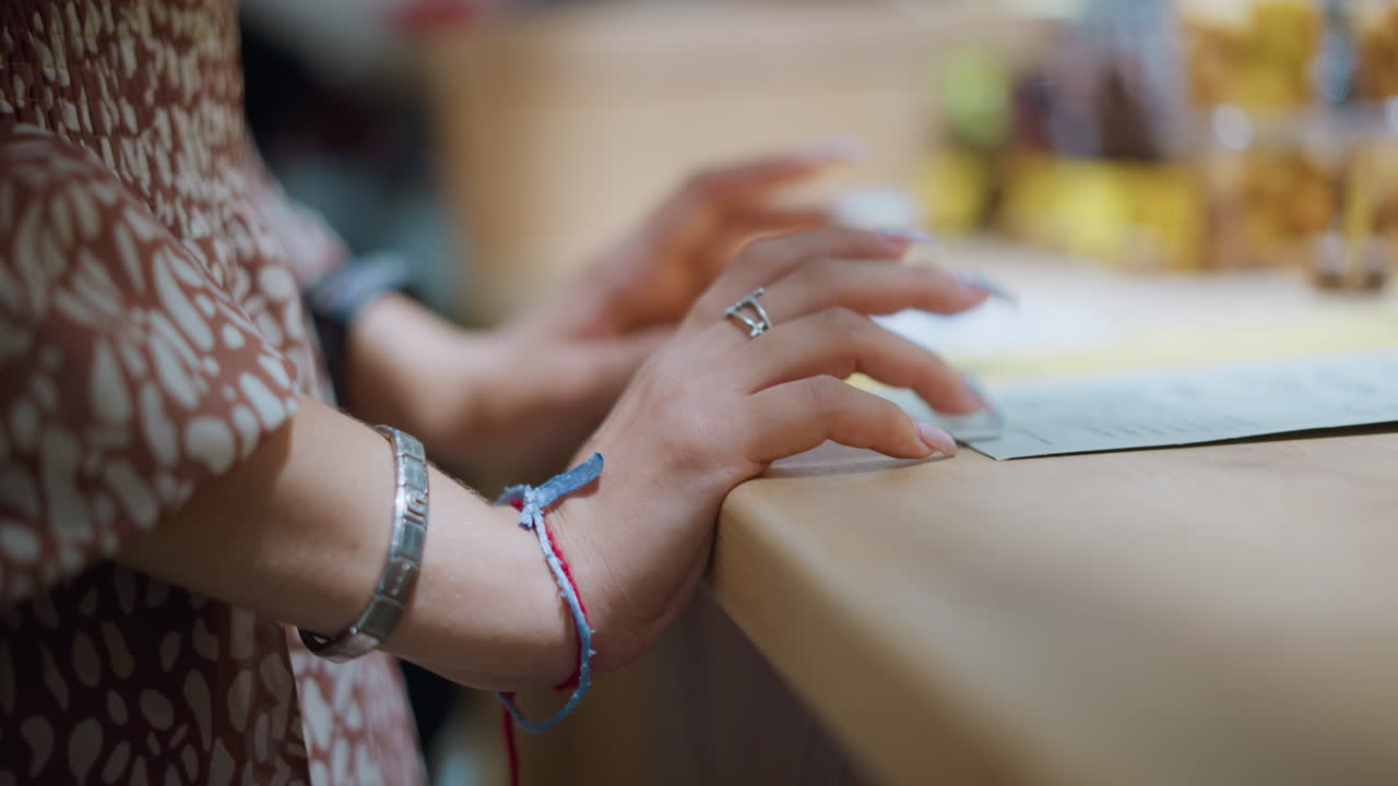 primer plano de las manos de la mujer adornadas con una pulsera de plata y un cordón rojo, uñas pulidas descansando suavemente sobre el papel en la mesa, sugiriendo contemplación o toma de decisiones, con un fondo suave y borroso