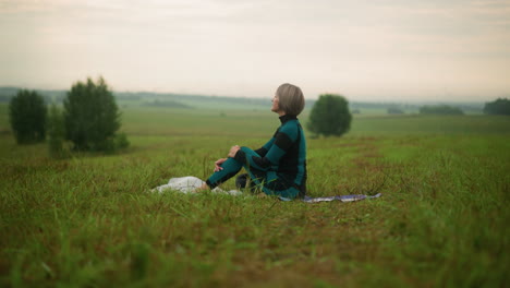 woman seated on yoga mat in misty grassy field, holding her knee, looking focused into the distance, surrounded by nature, she embraces calmness, engaging in peaceful mindfulness meditation