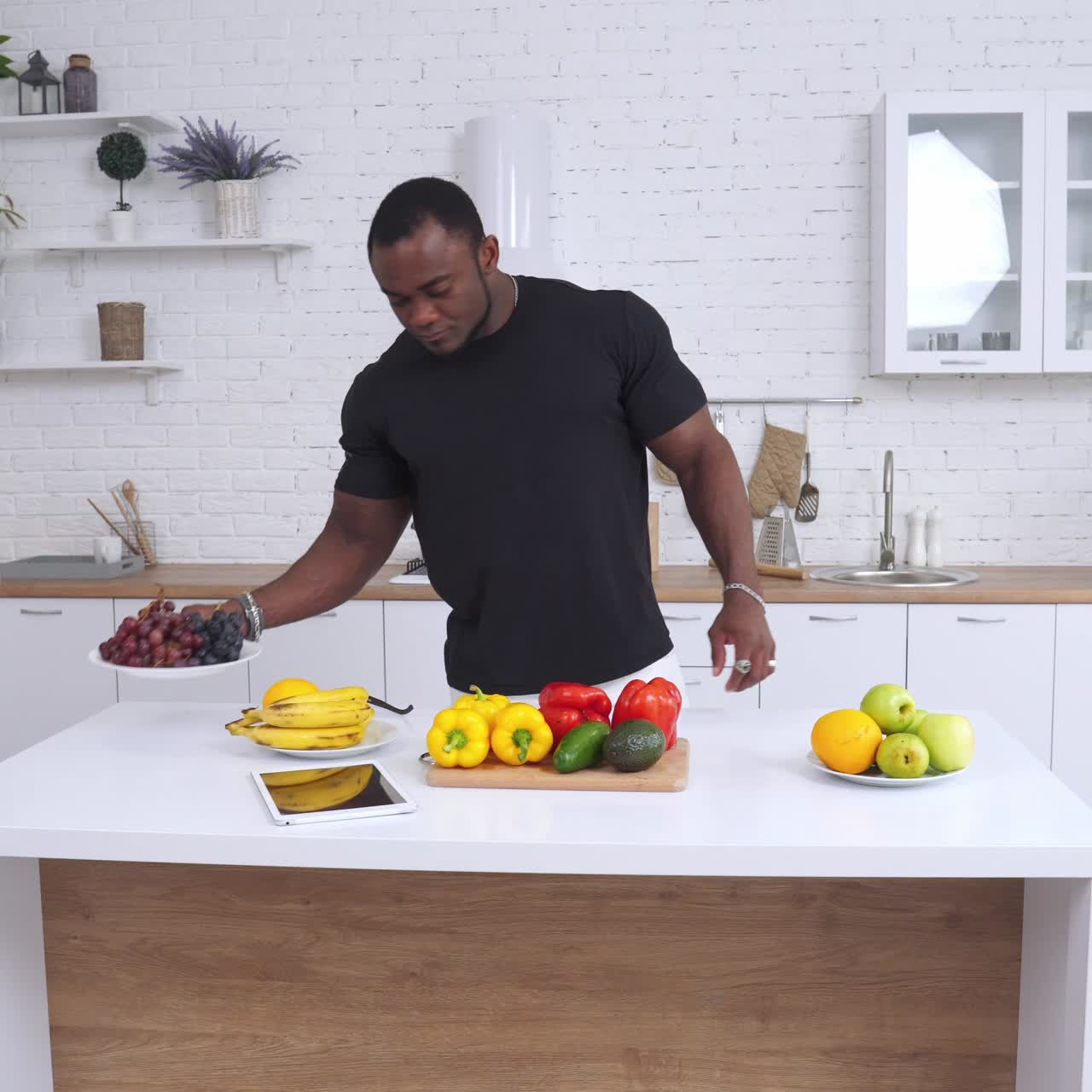 Un uomo afroamericano forte e muscoloso guarda il tavolo con molta frutta e verdura. Uno sportivo controlla il suo iPad per informazioni su una dieta sana