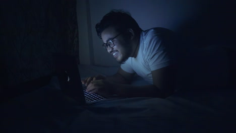 happy indian man using laptop at late night on bed, typing on laptop