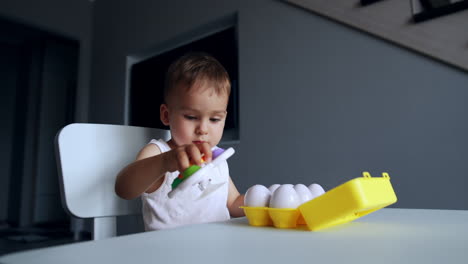 adorable toddler boy sits down at the desk. smiling kid takes a toy and bangs it by the desk.