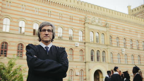 portrait of the gray-haired male professor in glasses and black gown crossing hands and smiling in front of the university