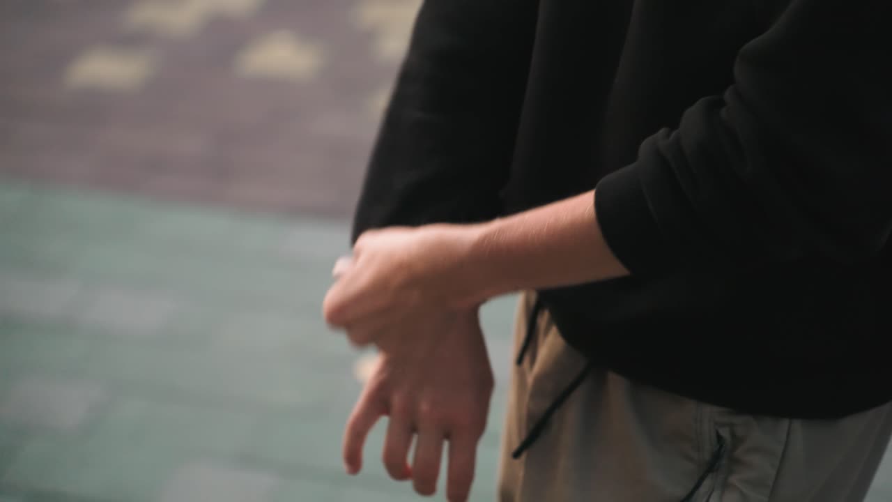 Primo piano di mani caucasiche, bagnate dalle gocce di pioggia, che si aggiustano la manica. Su una pavimentazione umida e con toni tenui, si intravedono i polsini arrotolati, l'avambraccio che si stabilizza, una presa rapida e un respiro regolare, forse di un pendolare o di un artista.