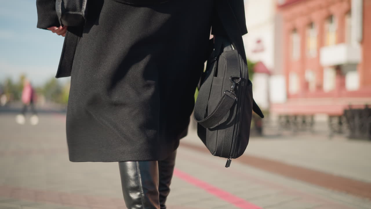 Back view of woman walking confidently on sunlit urban street, wearing chic black coat and stylish leather boots, carrying two bags, blurred background shows another person walking in pink jacket