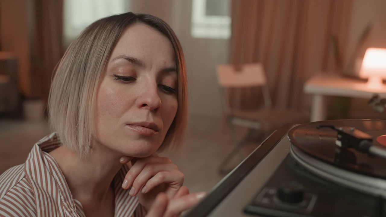 Woman enjoying vinyl records at home. Caucasian woman immersed in soulful music relaxation. Contemplative lady appreciating nostalgic melodies on her turntable illuminated by gentle warm glow