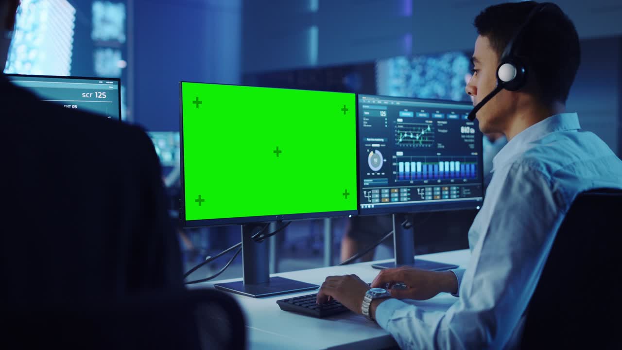 científico de datos masculino confiado trabaja en una computadora personal con plantilla de pantalla verde con auriculares en una gran sala de control y monitoreo. ingeniero joven en una sala de oficina de centro de llamadas con colegas.