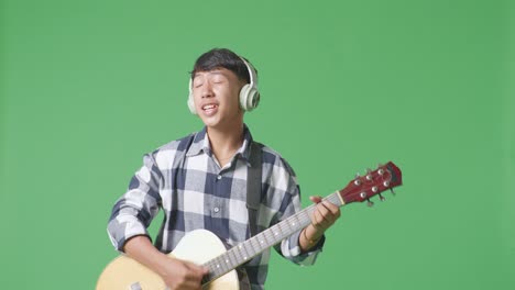 young asian teen boy wearing headphone playing guitar and singing on the green screen background