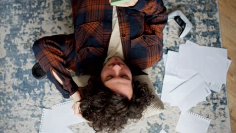 rotating camera happy brunette guy with curly hair lies on the floor on a pillow and types his ideas into a green smartphone