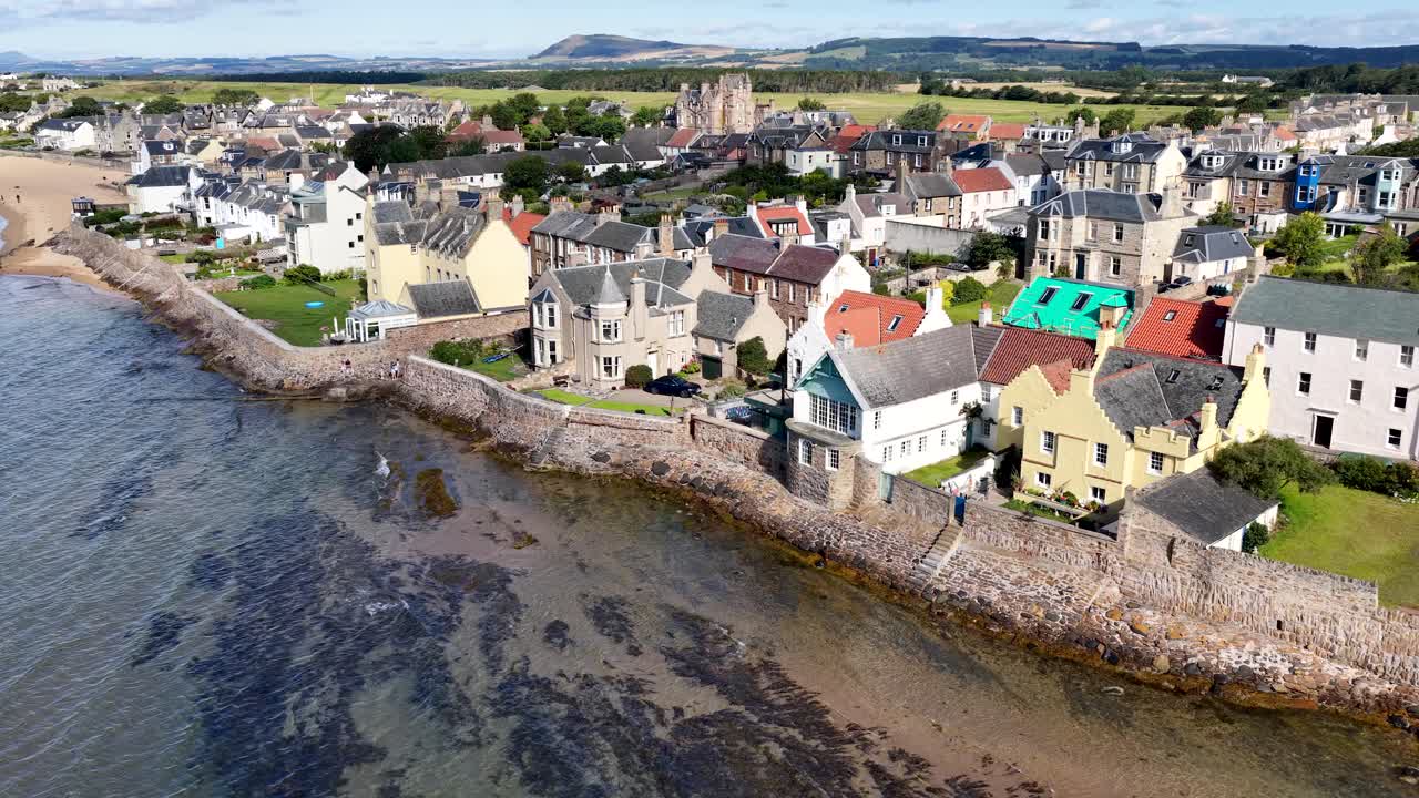 Drone glides above historic seaside village, stone houses, and shoreline under bright daylight