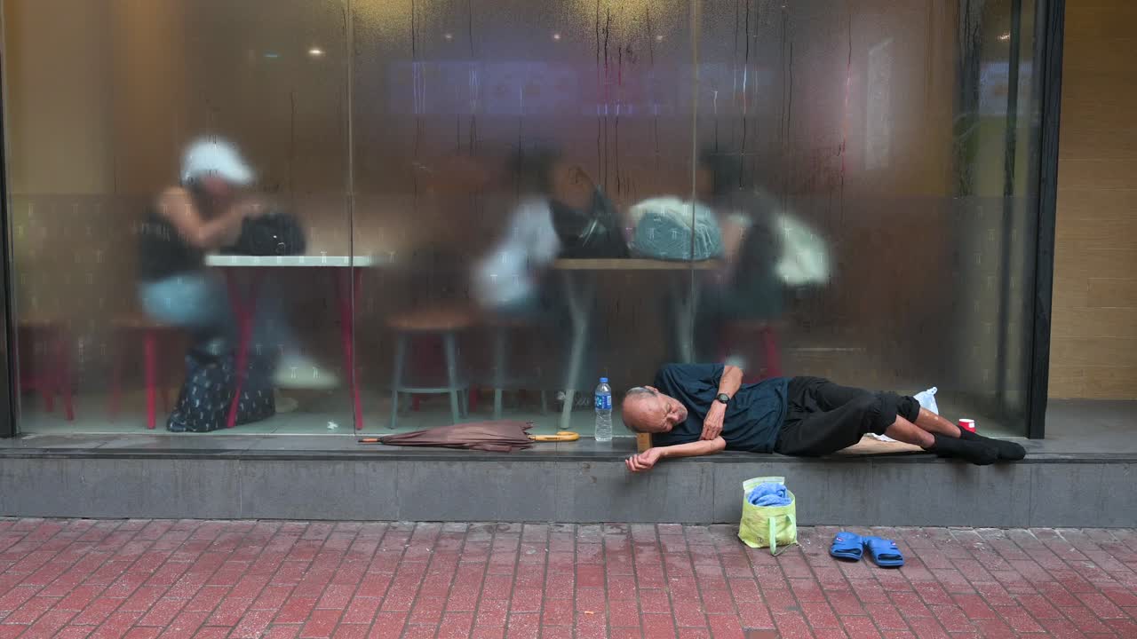 An elderly homeless man sleeps on the sidewalk in front of a restaurant, with patrons visible inside and pedestrians hurrying by on the streets of Hong Kong.