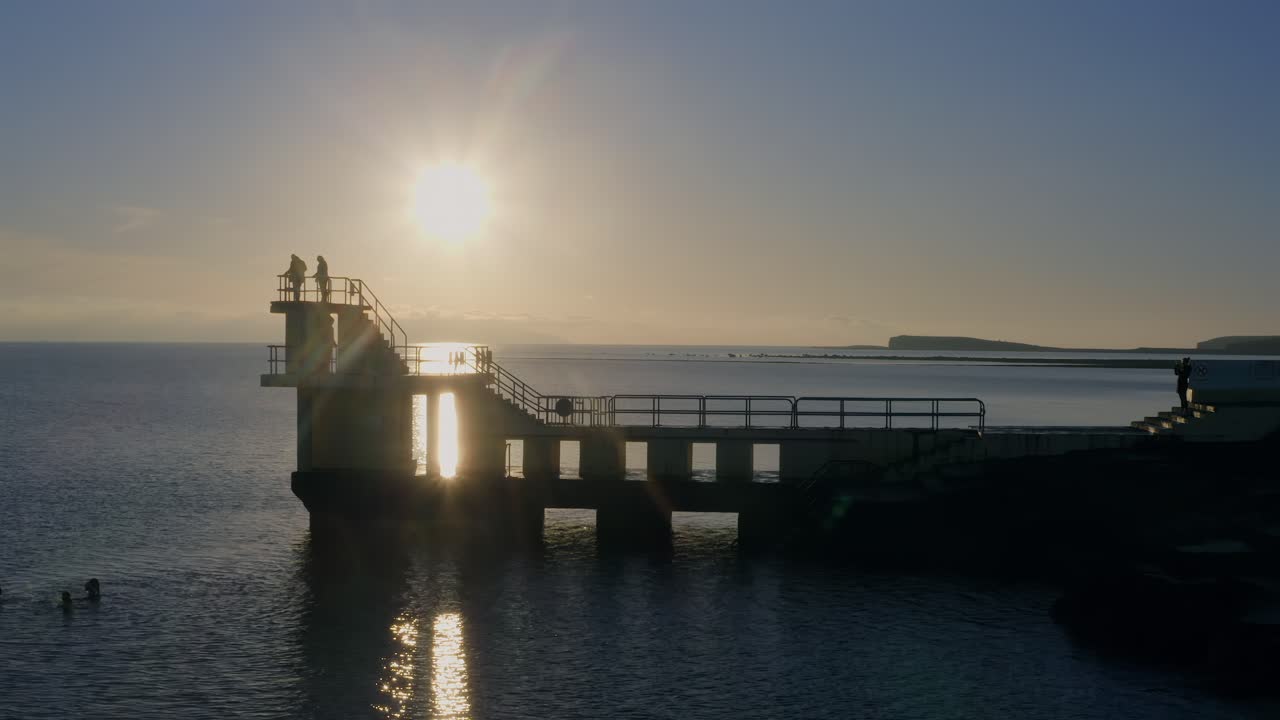 Aerial orbit of Blackrock diving board with silhouetted dippers and visitors at sunset. Galway
