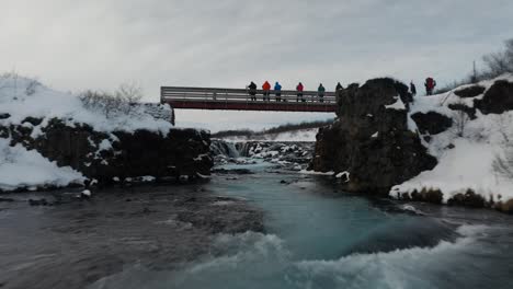 Wunderschöne-Drohnenaufnahme-Unter-Der-Brücke-Für-Den-Fluss,-Während-Sich-Menschen-Auf-Der-Brücke-Befinden-Und-Schnee-Alles-Bedeckt