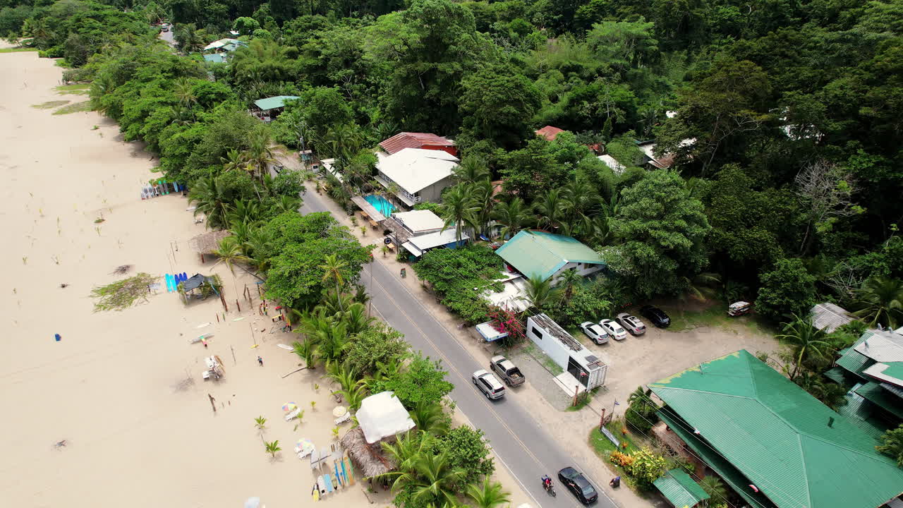 A dynamic aerial of a busy road along the popular beach Playa Cocles as tourists find surfboards and cars drive by the sand and palm tree lined coast of the Caribbean city of Puerto Viejo, Costa Rica