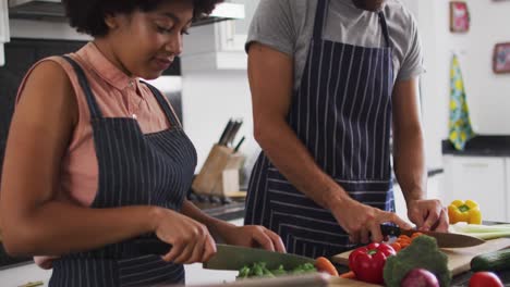 Mixed-race-couple-wearing-aprons-chopping-vegetables-together-in-the-kitchen-at-home