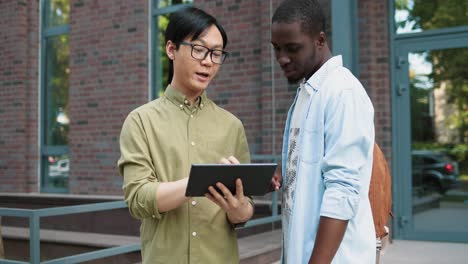 Camera-zooming-on-asian-man-talking-with-his-caucasian-african-american-classmate-watching-something-on-the-tablet-and-talking-in-the-street-near-the-college