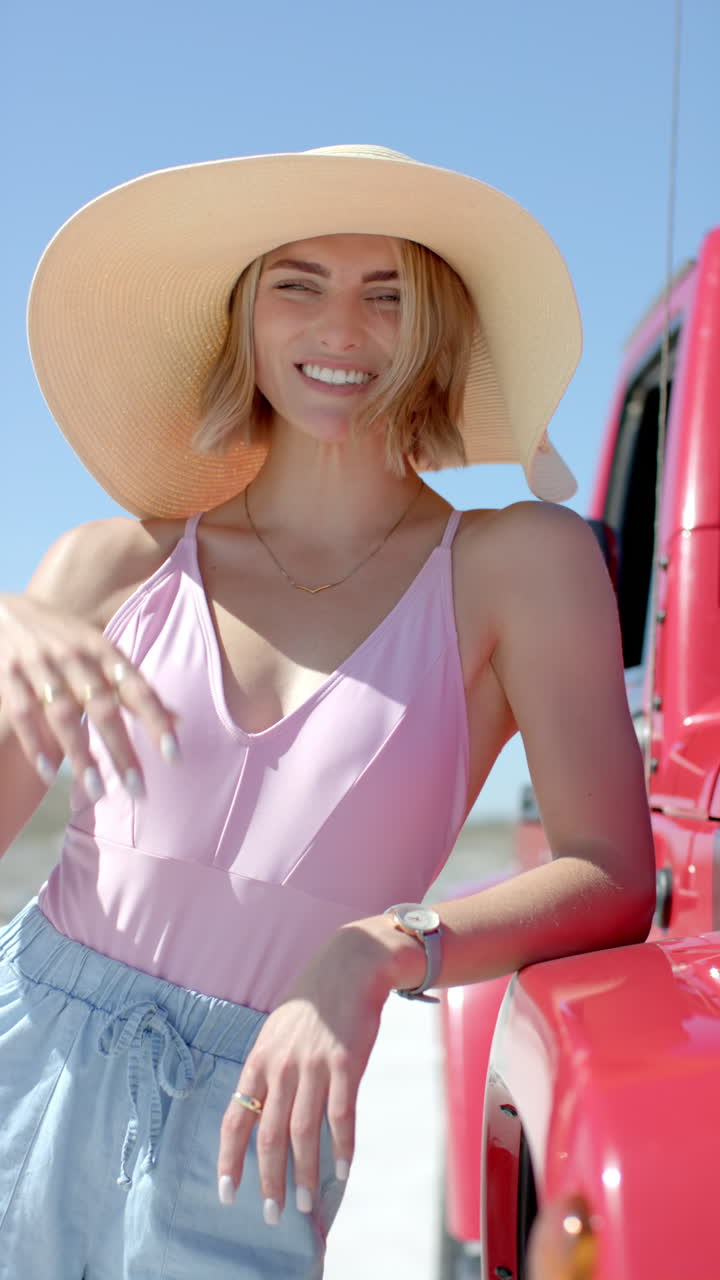 Vertical video: Smiling woman in sunhat leaning on red car during road trip on sunny day