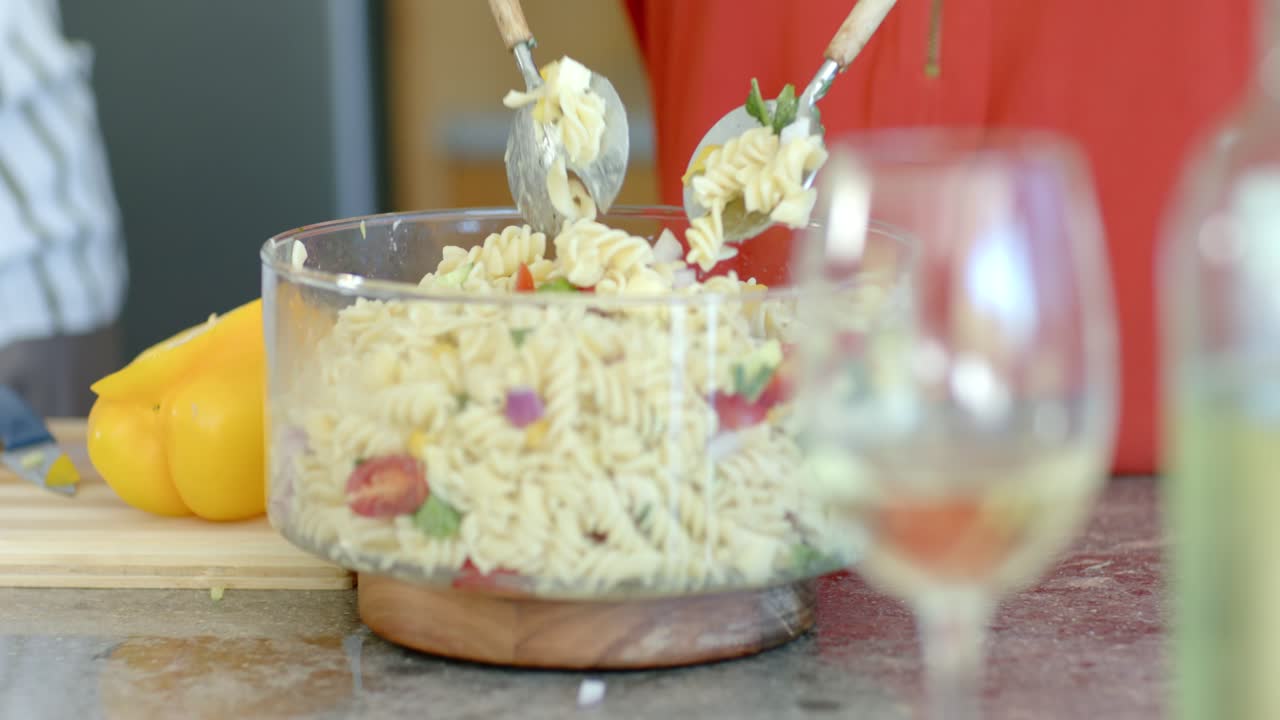 Concentrating and adjusting grip, cook's hands are stirring rotini salad in glass bowl to combine