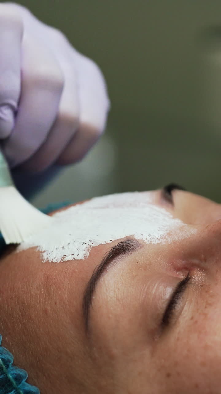 Beautician in latex gloves uses brush to cover client's face with mask. Rejuvenation procedures in beauty salon. Close up. Vertical video