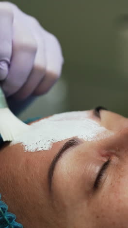 beautician in latex gloves uses brush to cover client's face with mask. rejuvenation procedures in beauty salon. close up. vertical video