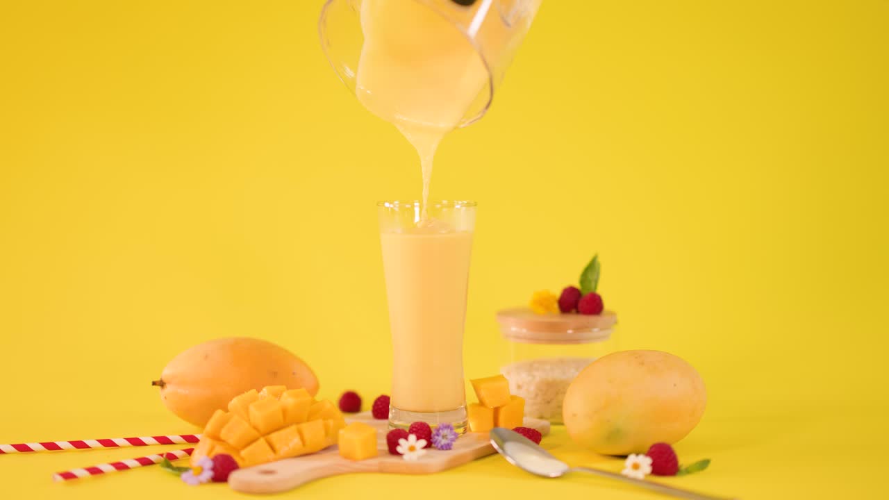 Hand pours thick mango smoothie into tall glass among tropical fruit on yellow backdrop