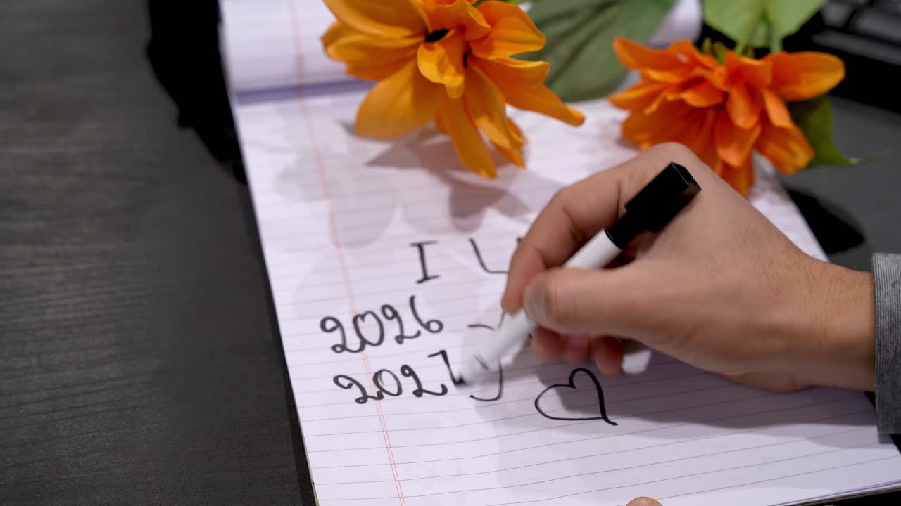 Primo piano di una mano che scrive un biglietto "Ti amo" con delle date su carta a righe, fiori arancioni nelle vicinanze su una scrivania scura, illuminazione interna soffusa con profondità di campo ridotta.