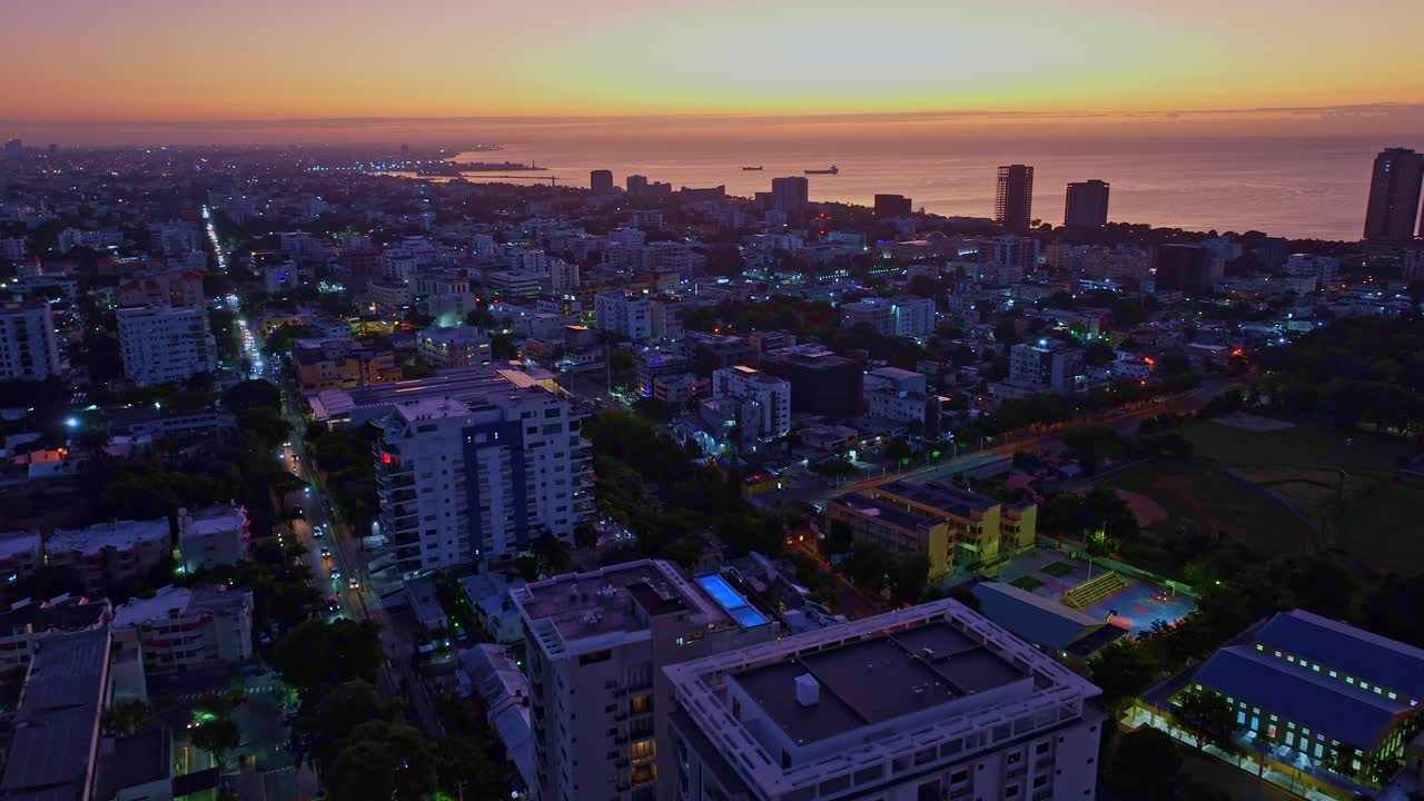 Forward aerial shot of Santo Domingo at sunrise, showcasing the skyline, Caribbean Sea, and golden sunlight casting warm hues over the buildings and horizon in a serene morning scene