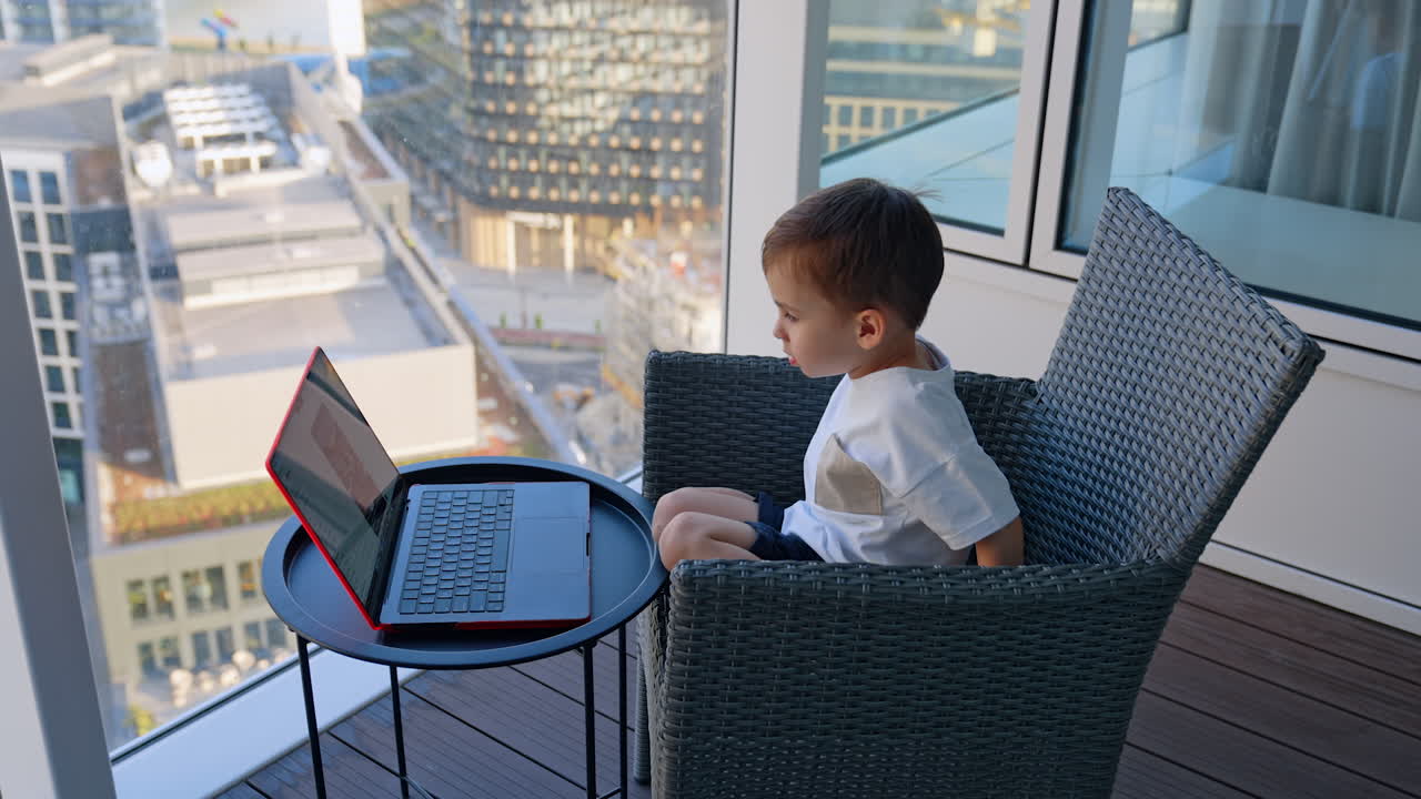 Cute little boy sits in the armchair concentrated on laptop. Focused kid watches video at home. High-angle view