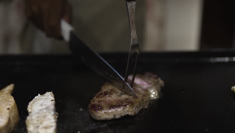 chef cutting, slicing medium rare beef steak sarlion into peaces while cooking on black satake frying table in japanese restaurant - hands close-up slow motion