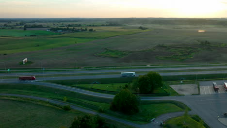 vista aérea de una carretera rural con camiones viajando, rodeada de vastos campos verdes y marrones, con un pequeño pueblo visible en el fondo