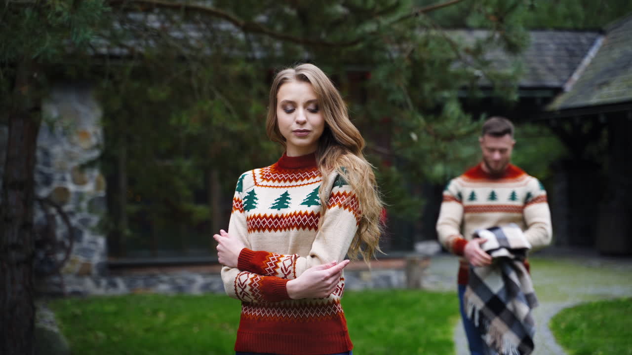 Lovely girl and a guy outdoors. Man covering his girlfriend's shoulders with a blanket in autumn in the yard. Romantic couple in warm sweaters in terrace.