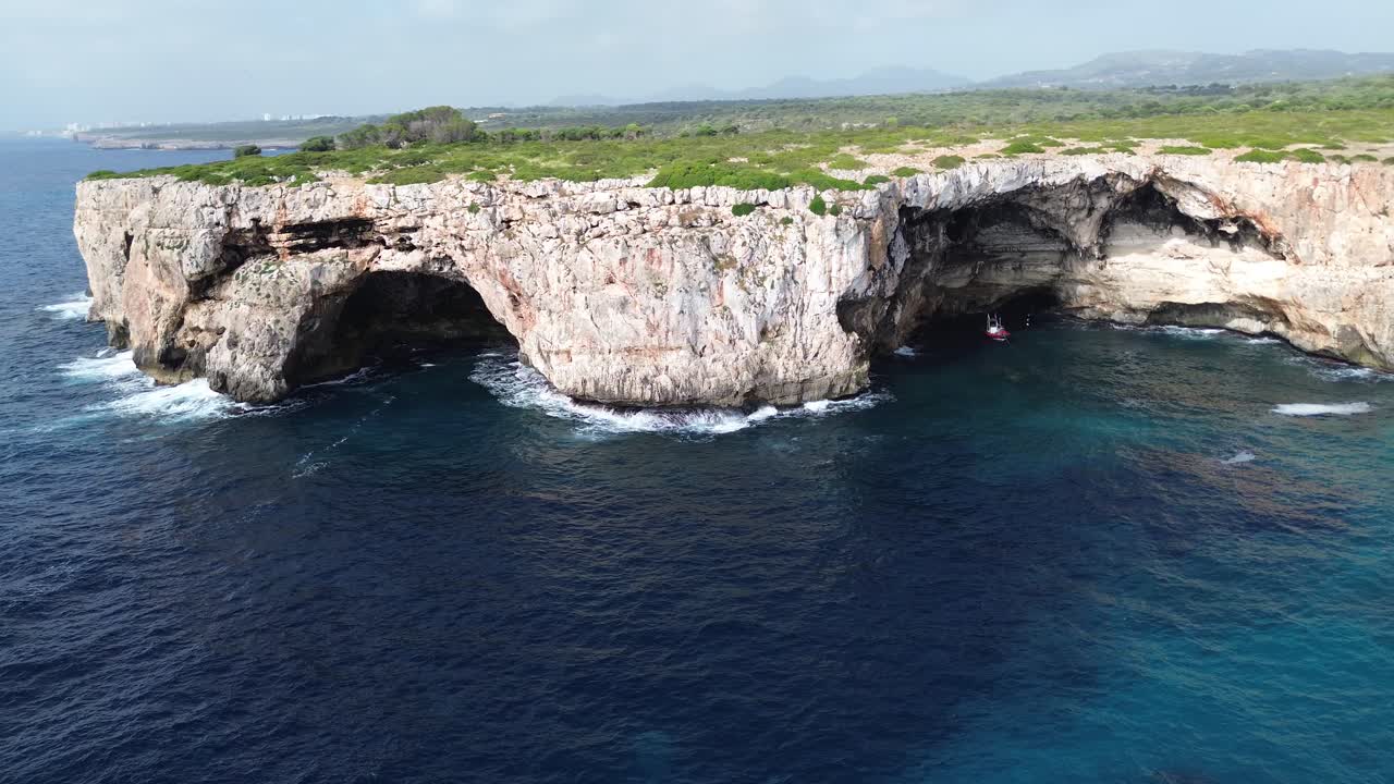 Dynamic 4K drone footage arching sideways around a rocky shoreline in Mallorca. Features crystal clear water, cliffs, green trees, and a red dive boat inside a large sea cave. Captured June 2025